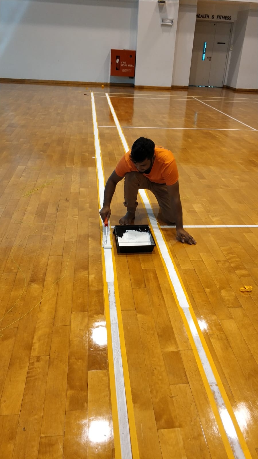 Court marking work in progress: white line paint applied between yellow boundary lines on a timber sports hall floor